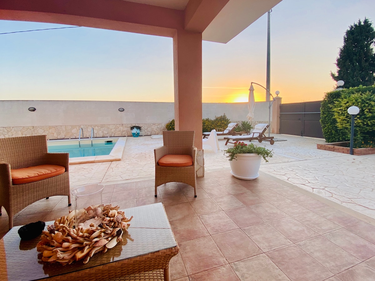 An outdoor seating area is visible, featuring two wicker chairs with orange cushions. A small table holds a decorative bowl filled with shells. In the background, a private pool is framed by a serene sunset, with lounge chairs positioned nearby and greenery accentuating the space.