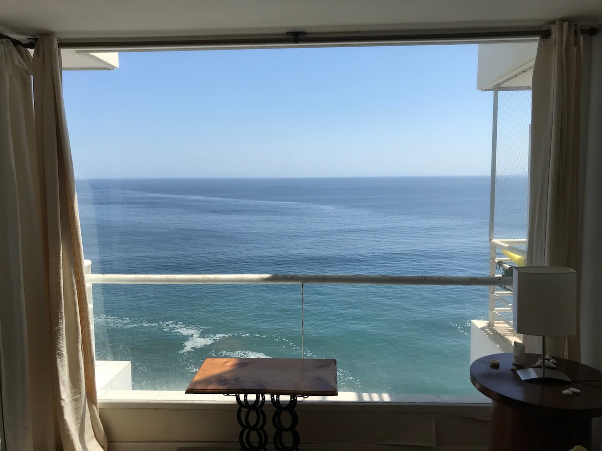 A large window is framed by light curtains, offering an unobstructed view of the calm ocean. A wooden table is positioned in front of the window, complementing the serene coastal scenery visible beyond.
