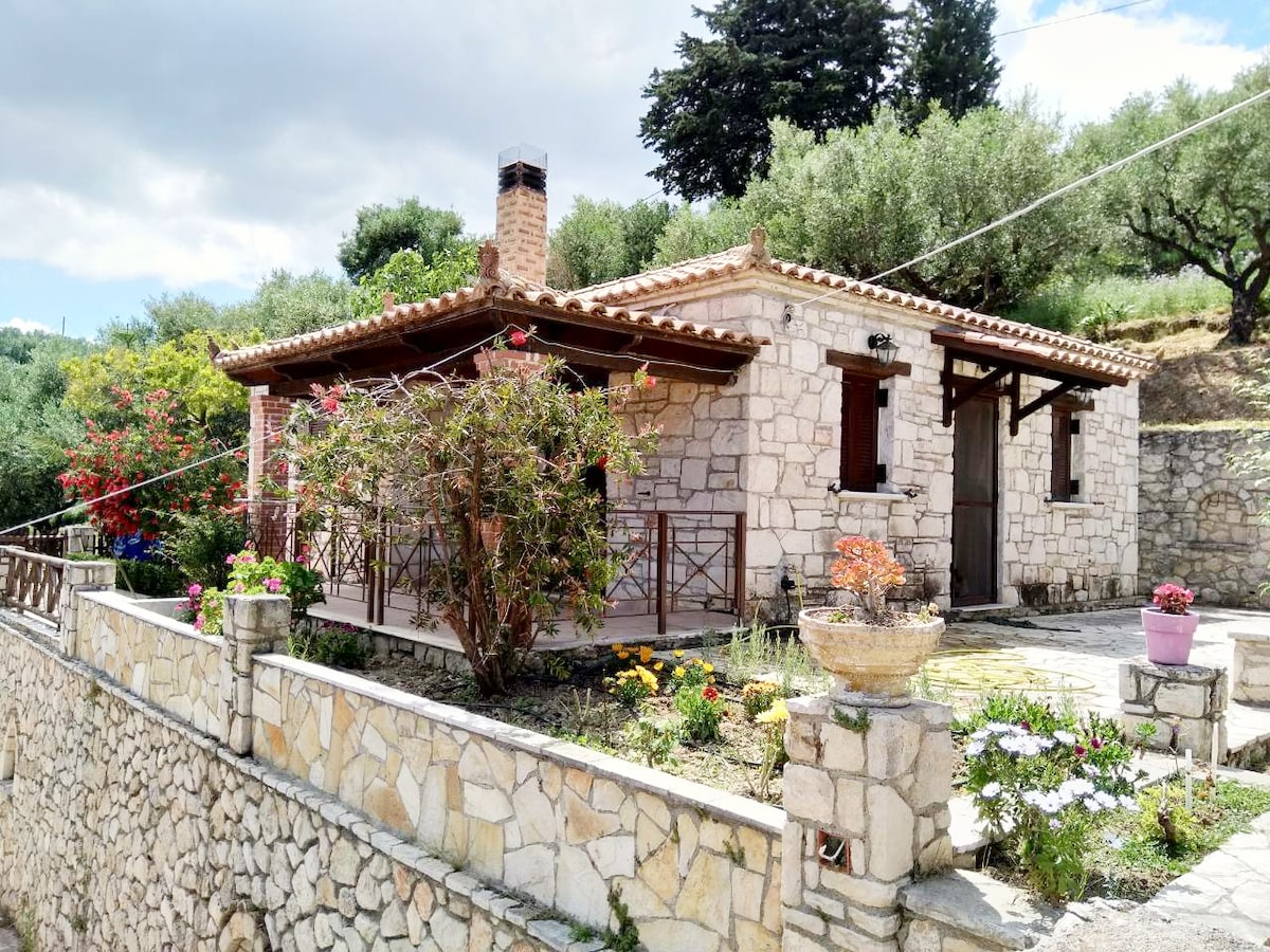 A traditional stone house is set amidst greenery, featuring a prominent porch with wooden railings. The exterior is adorned with vibrant flowers, with olive trees in the background. A chimney is visible on the roof, and the surrounding area is neatly landscaped.