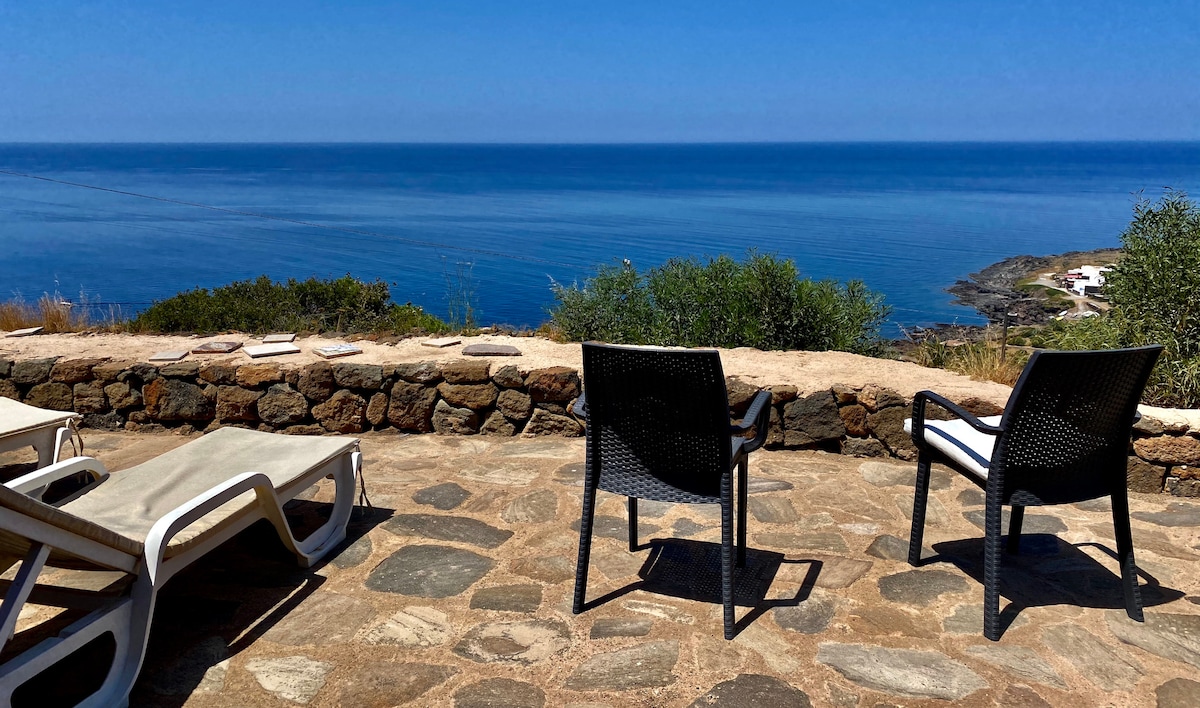 An outdoor terrace features two lounge chairs and a stone wall, providing unobstructed views of the calm sea. The landscape includes various green shrubs and rocky coastline, with a clear blue sky above, creating a serene setting for relaxation.