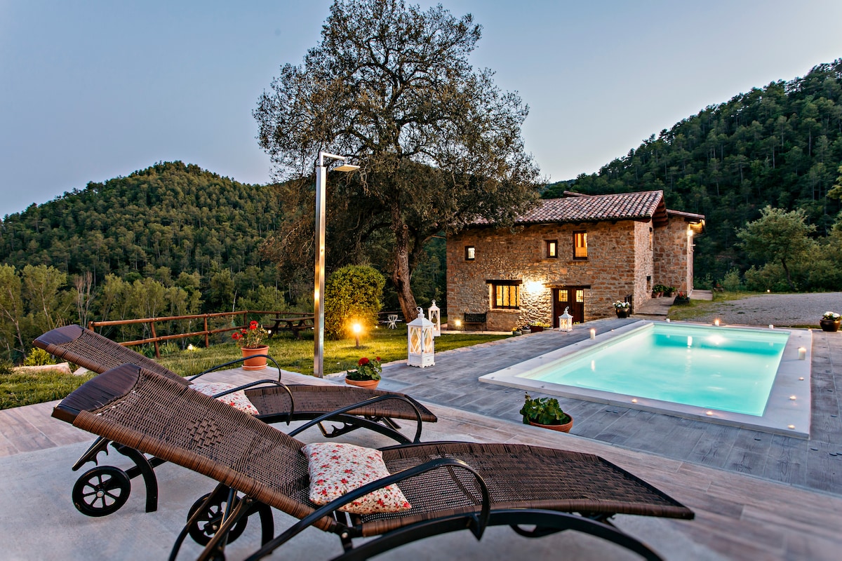 The image features an outdoor area with two lounge chairs positioned by a serene pool, surrounded by natural greenery. A rustic stone house with warm lighting is visible in the background, nestled against a mountainous landscape, providing a tranquil rural atmosphere.