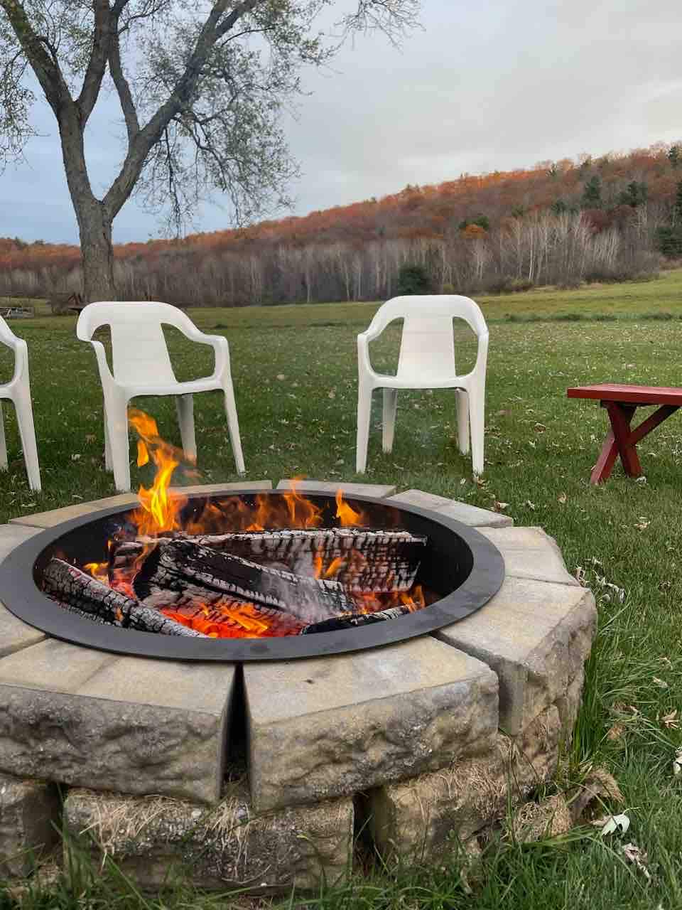A circular stone fire pit is surrounded by several white plastic chairs, providing a cozy seating area. The fire is burning brightly, with flames flickering above a bed of logs. In the background, trees show signs of autumn colors, and a grassy field stretches out.