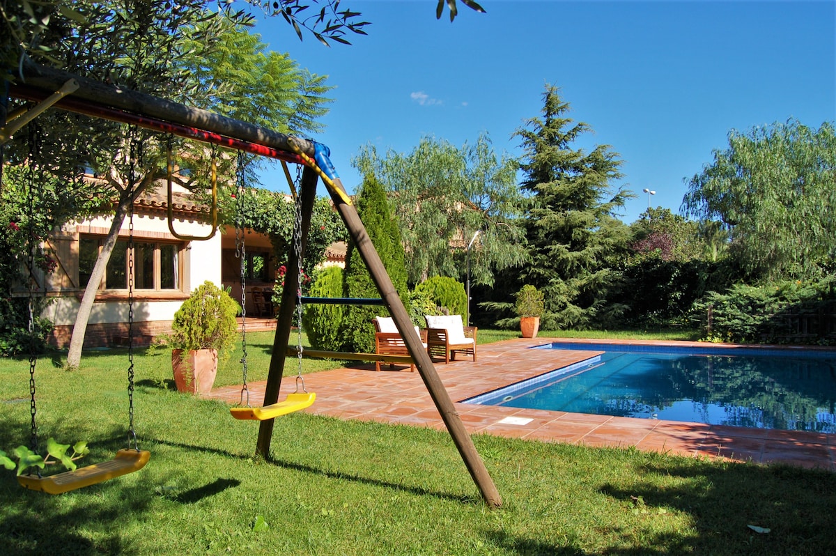 A private outdoor area is featured, showcasing a tranquil swimming pool surrounded by lush greenery. A wooden swing set is visible in the foreground, while comfortable seating arrangements are positioned near the pool. Potted plants add natural elements to the serene setting.