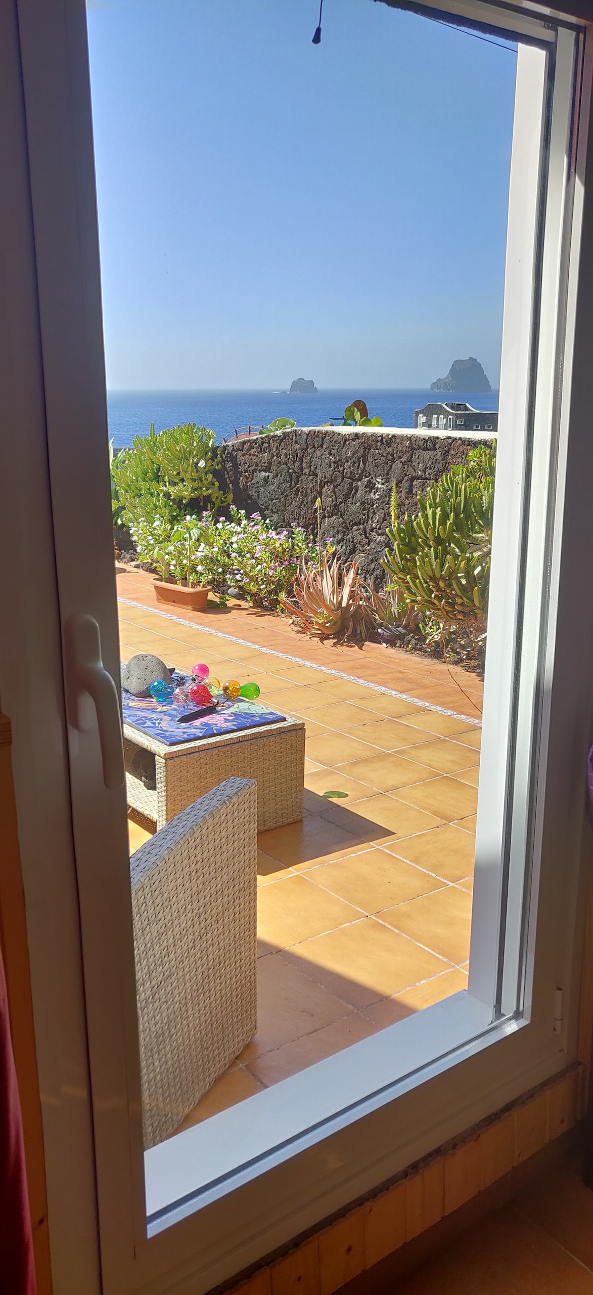 A view through a glass door reveals a charming outdoor seating area with a small table. Surrounding flowers and greenery frame the space, while the serene sea and distant rocky formations are visible in the background, creating a peaceful setting.