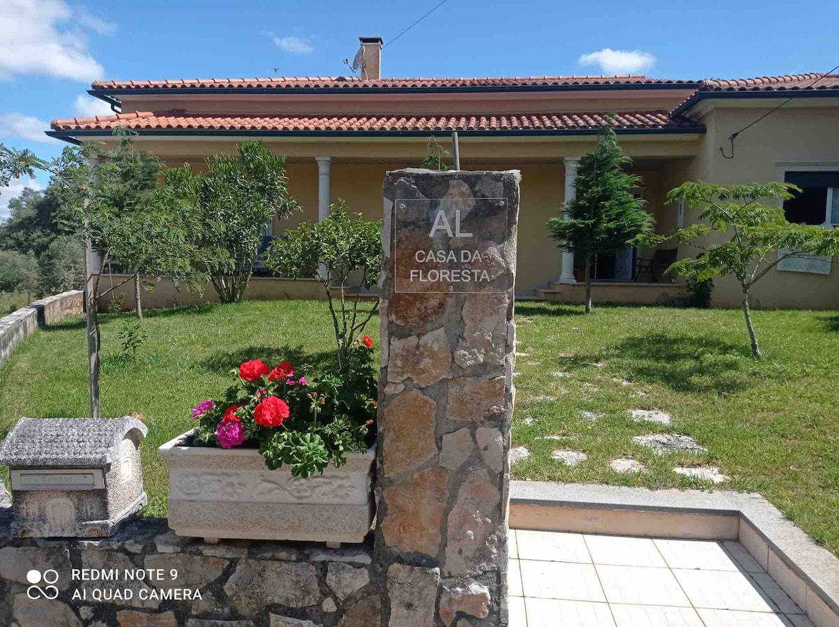 A simple stone entrance marker displays the name 'Casa da Floresta.' Colorful flower pots with blooming flowers add vibrancy to the entrance. The well-maintained front yard features a smooth pathway leading to the house, surrounded by grassy areas and a few small trees.