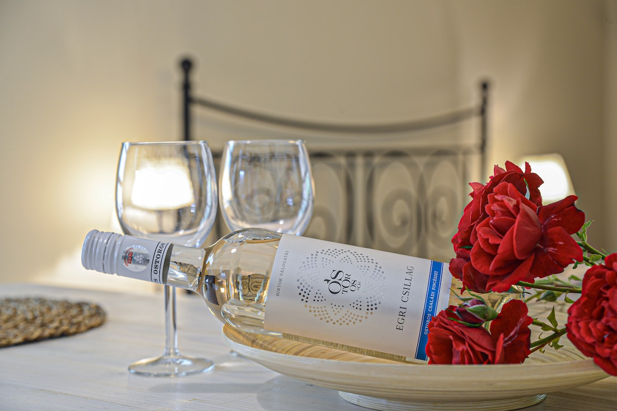 A bottle of Egri Csillag wine is placed in a shallow dish, accompanied by two glasses and a bouquet of red roses. Soft lighting from two lamps creates a relaxing atmosphere, highlighting the elegant simplicity of the arrangement.