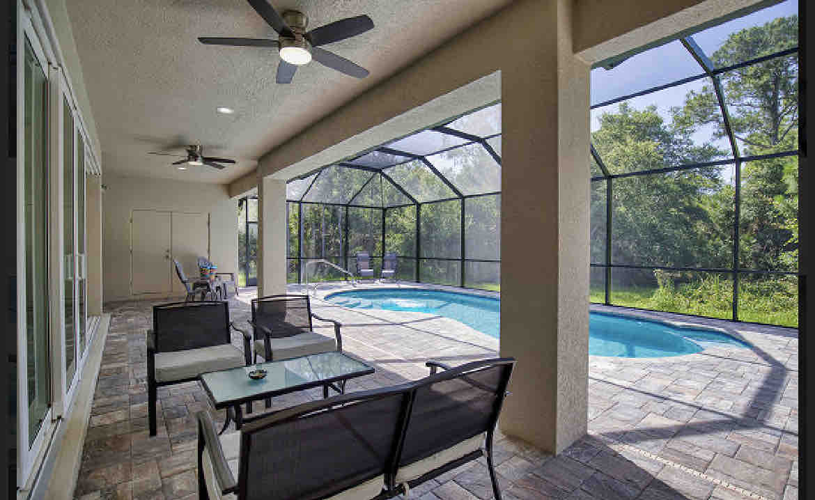 The enclosed lanai features a seating area with comfortable chairs and a table, overlooking a private pool surrounded by lush greenery. Ceiling fans provide airflow, and large screens create a seamless outdoor experience, inviting relaxation and leisure.