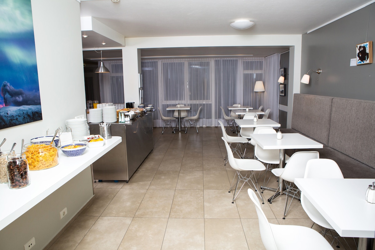 A communal kitchen area is shown, featuring white table settings with modern chairs. An assortment of food items is displayed on a counter alongside a stainless-steel serving area. Natural light fills the room through sheer curtains, creating a bright and open atmosphere.