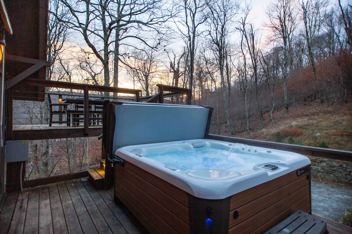 A hot tub is positioned on a wooden deck surrounded by trees, creating a serene outdoor setting. The hot tub features a paneled exterior and is ready for use, while the landscape reflects the soft colors of dusk.