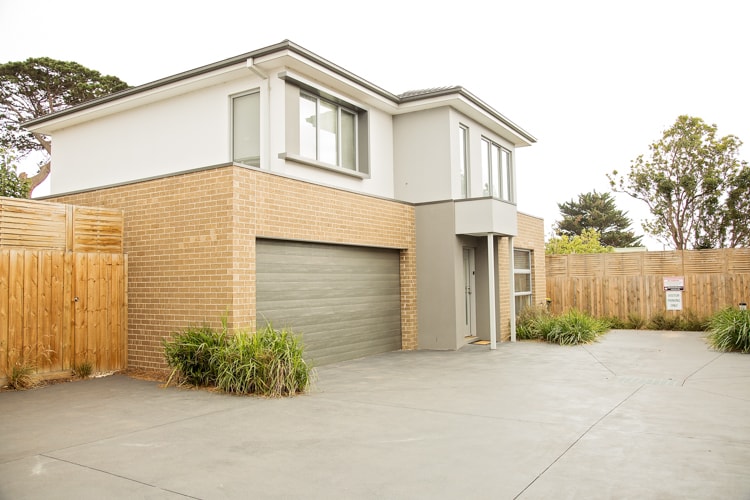 The exterior of the townhouse is displayed, featuring a modern two-story design with a blend of brick and smooth finishes. A spacious driveway leads to a garage, framed by landscaped greenery. Large windows allow for natural light, and wooden fencing provides privacy.