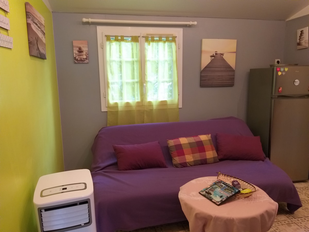 A living space features a purple sofa with cushions and a small round table. Natural light enters through windows adorned with sheer green curtains, brightening the room. Wall art depicting serene landscapes contributes to a calming ambiance. A compact fridge is also visible in the background.