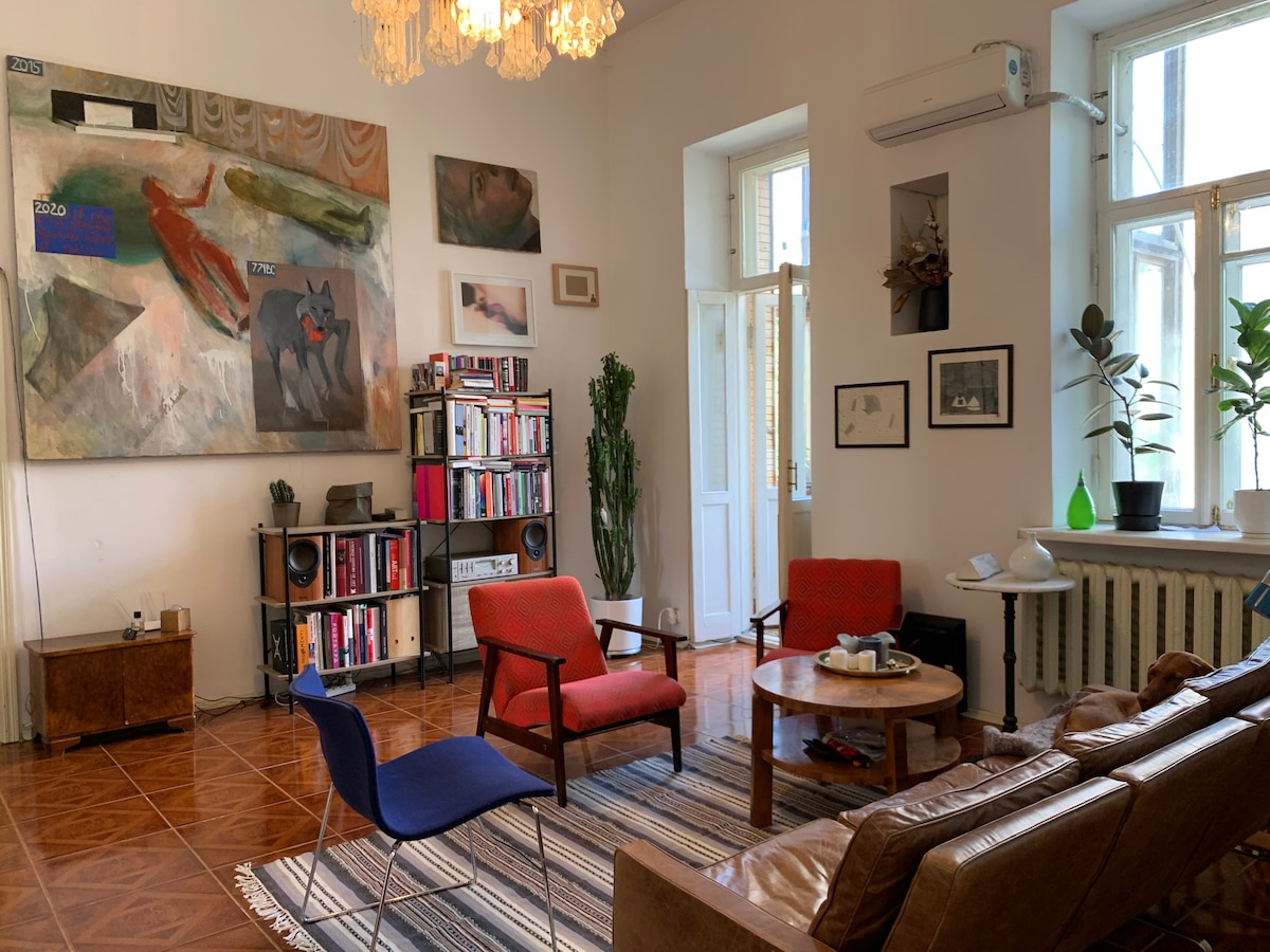 A spacious lounge area is highlighted by large windows allowing natural light. Colorful chairs sit around a circular table, and a vintage sofa complements the atmosphere. An artwork decorates the wall, complemented by shelves of books. Potted plants add greenery to the space.