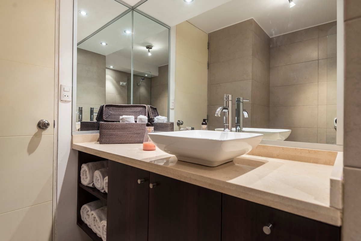 A well-appointed bathroom features a modern sink atop a beige countertop with a spacious mirror above. Neatly rolled towels are stored in a cabinet beneath, while an elegant soap dispenser is placed beside the sink. Soft lighting enhances the serene atmosphere.