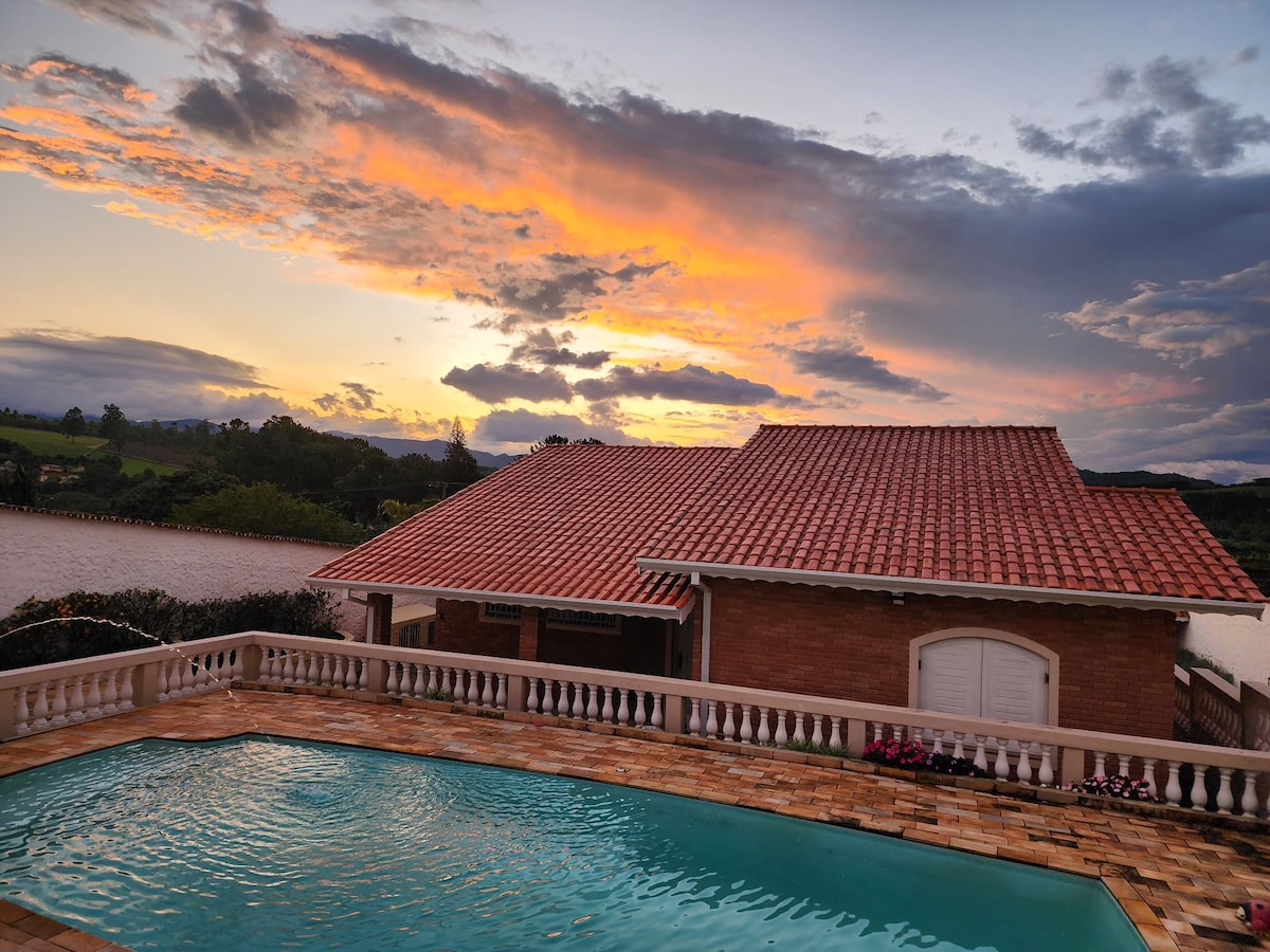 A serene sunset casts warm hues over a spacious outdoor area, featuring a sparkling pool. The house, with its terracotta roof, sits adjacent to greenery and water, providing a tranquil atmosphere. Colorful flowers line the balcony railing, enhancing the scene.