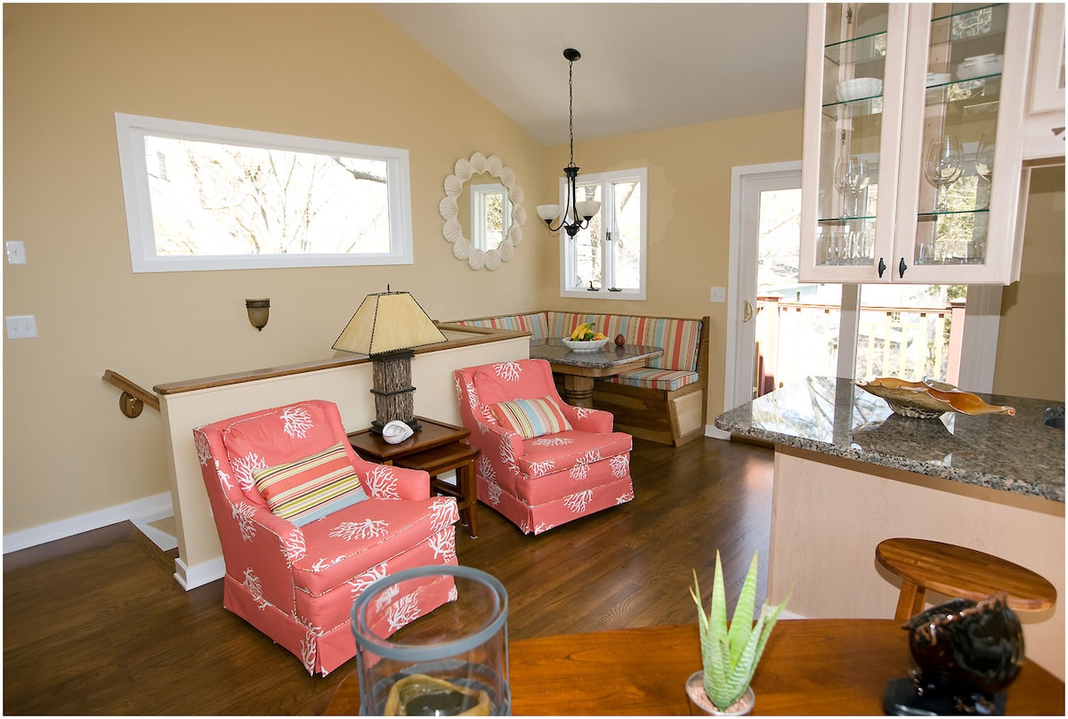 A bright living area features two coral-colored armchairs with floral patterns, a round mirror on the wall, and a small dining nook with striped seating. Natural light floods in through multiple windows, enhancing the warm tones of the wooden flooring and kitchen cabinetry.