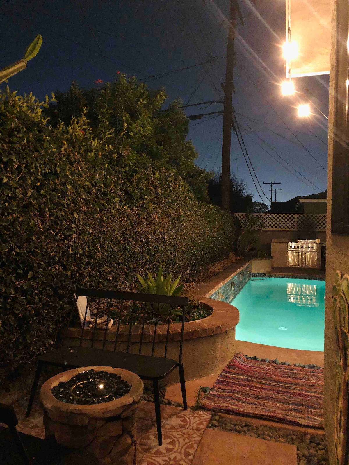 An outdoor seating area is illuminated by soft string lights, creating a cozy atmosphere. A small black chair is positioned near the edge of a swimming pool surrounded by lush greenery. The pool water reflects the night sky, providing a tranquil setting.