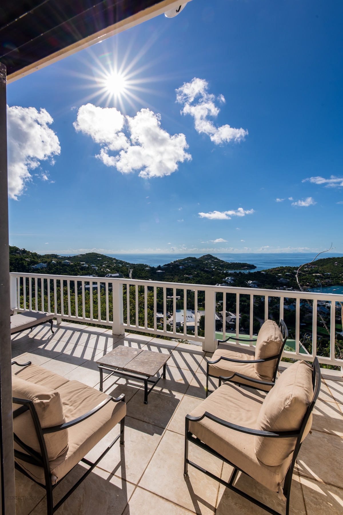 A spacious outdoor deck features comfortable seating with beige cushions arranged around a square table. The sun shines brightly overhead, illuminating the expansive view of the surrounding landscape and distant waters, providing a serene setting for relaxation.