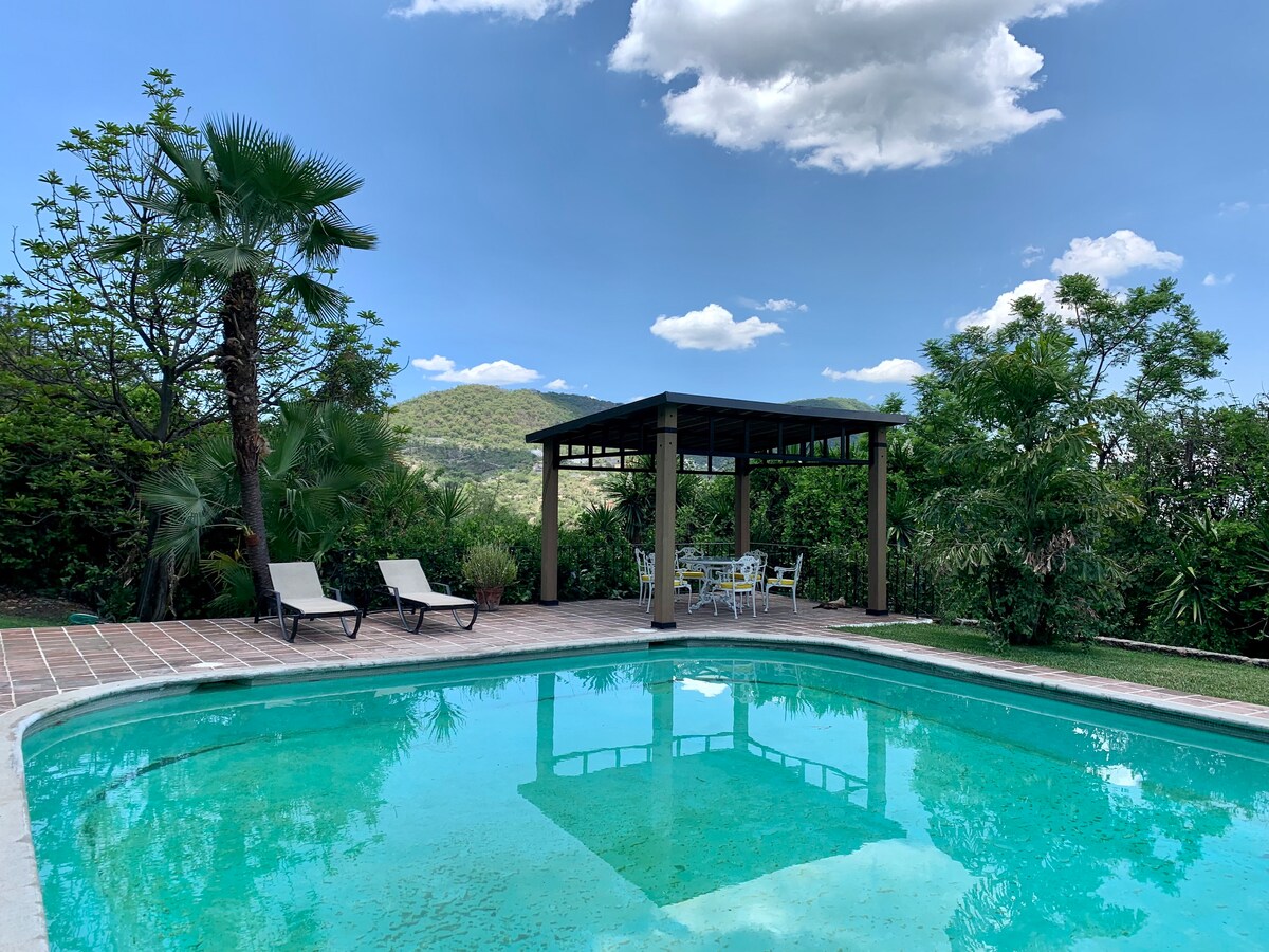 A refreshing swimming pool is situated outdoors, surrounded by lush greenery. Sun loungers are placed beside the pool, while a shaded seating area with a table and chairs is visible in the background, offering a place to relax and enjoy the mountain views.