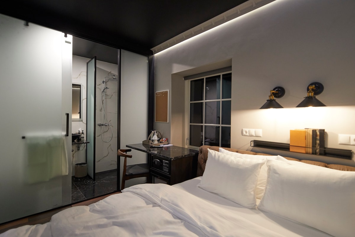 A compact room showcases a tailor-made luxury bed with crisp white linens. An ensuite bathroom is visible through frosted glass doors, featuring a rainfall shower. A work desk with a tea kettle and minibar is positioned nearby, while warm lighting highlights the stylish decor.