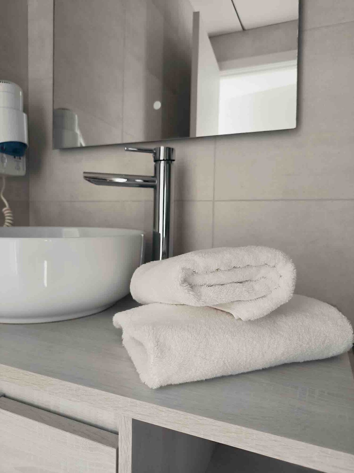 A clean, modern bathroom features a round basin on a minimalistic countertop. Two neatly rolled towels are placed beside the basin. A wall-mounted mirror reflects the soft, neutral tones of the tiles, enhancing the overall bright and airy feel.