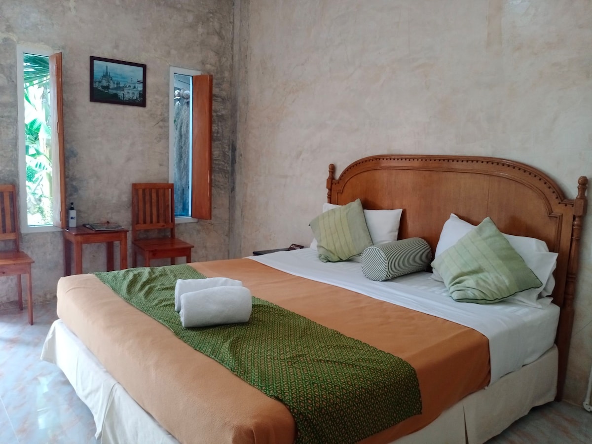 A spacious bedroom features a large wooden bed with decorative pillows and a rolled towel. Two wooden chairs and small tables are positioned near the windows, which offer natural light and views of greenery. The walls are finished in a light, textured material.