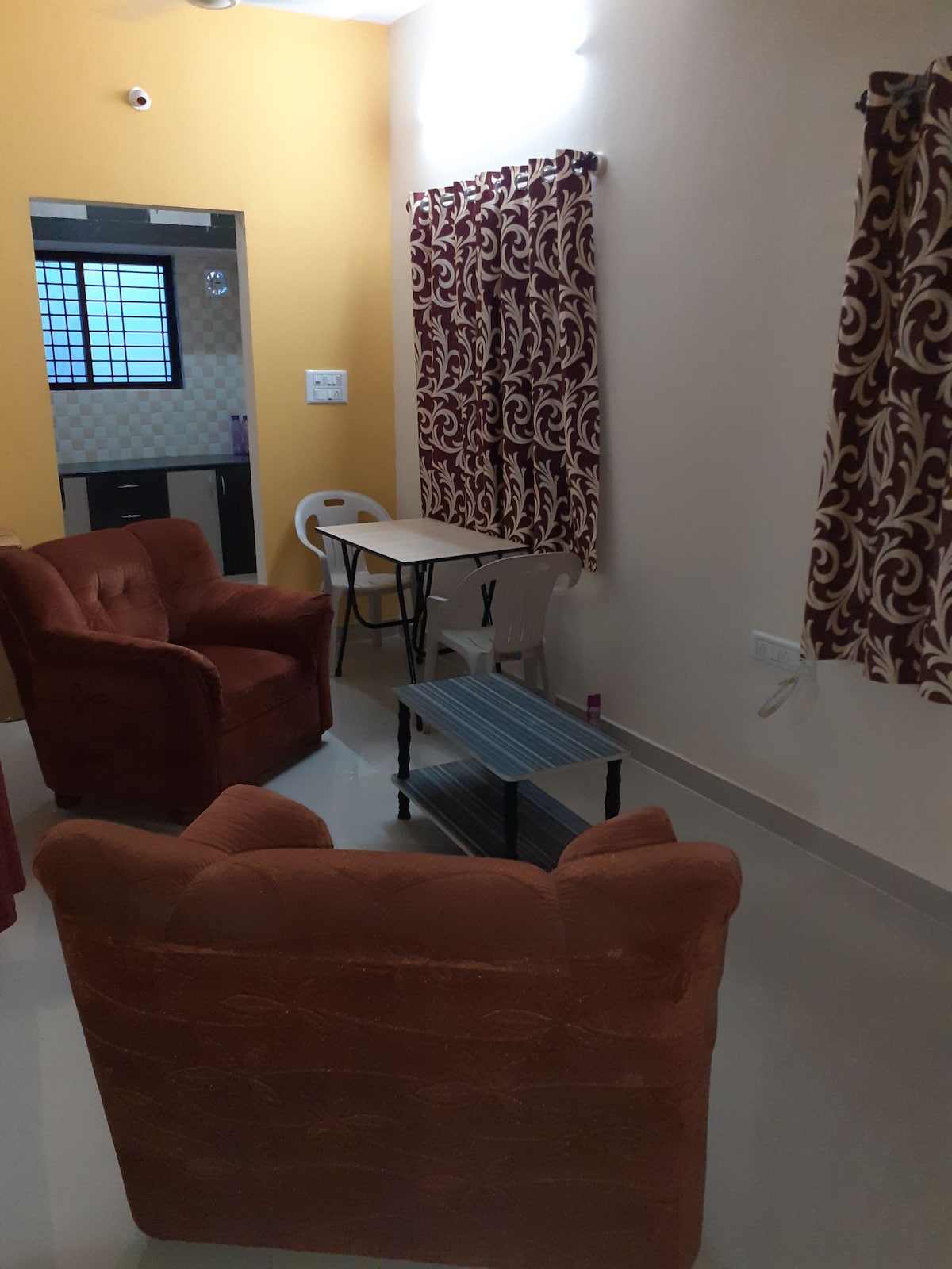 A cozy seating area features two plush armchairs with a small coffee table in between. A dining table with chairs is visible in the background, alongside a kitchen area partially enclosed by a partition. Natural light enters through a window covered by patterned curtains.