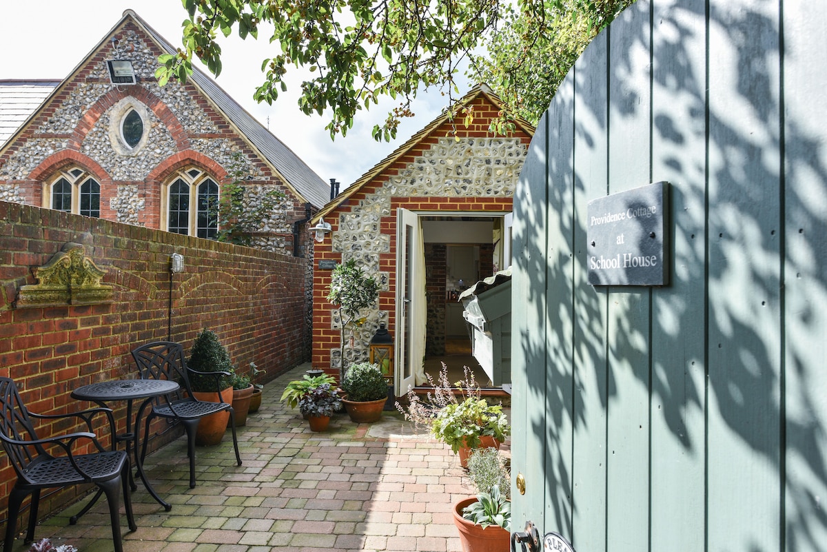 The entrance to Providence is framed by a rustic wooden gate and paved courtyard, showcasing a mix of potted plants and small seating areas with wrought iron chairs and a table. The adjacent building features traditional brick and flint, enhancing the charming atmosphere.