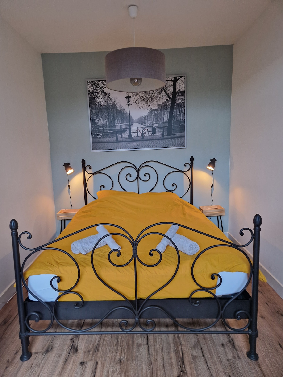 A cozy sleeping area features a black metal bed frame with an intricate design. A bright yellow blanket covers the spacious bed, complemented by neatly arranged towels. Soft bedside lamps provide gentle illumination, while a large framed photograph of a canal scene adds a touch of charm to the wall.
