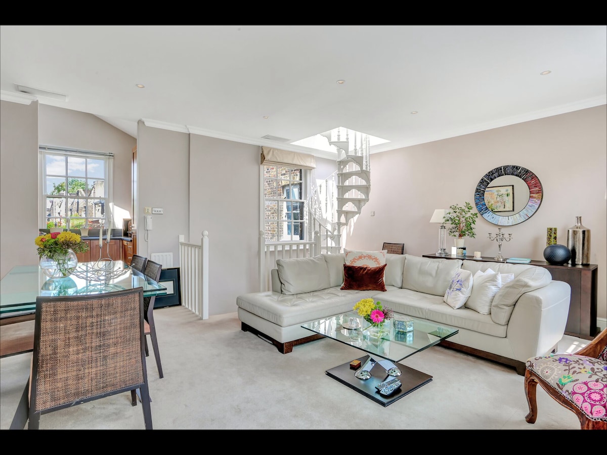 A bright and spacious living area features a large white sectional sofa adorned with decorative pillows. A glass coffee table sits atop a neutral carpet, surrounded by sleek dining chairs. Natural light floods through the windows, highlighting the elegant spiral staircase in the background.