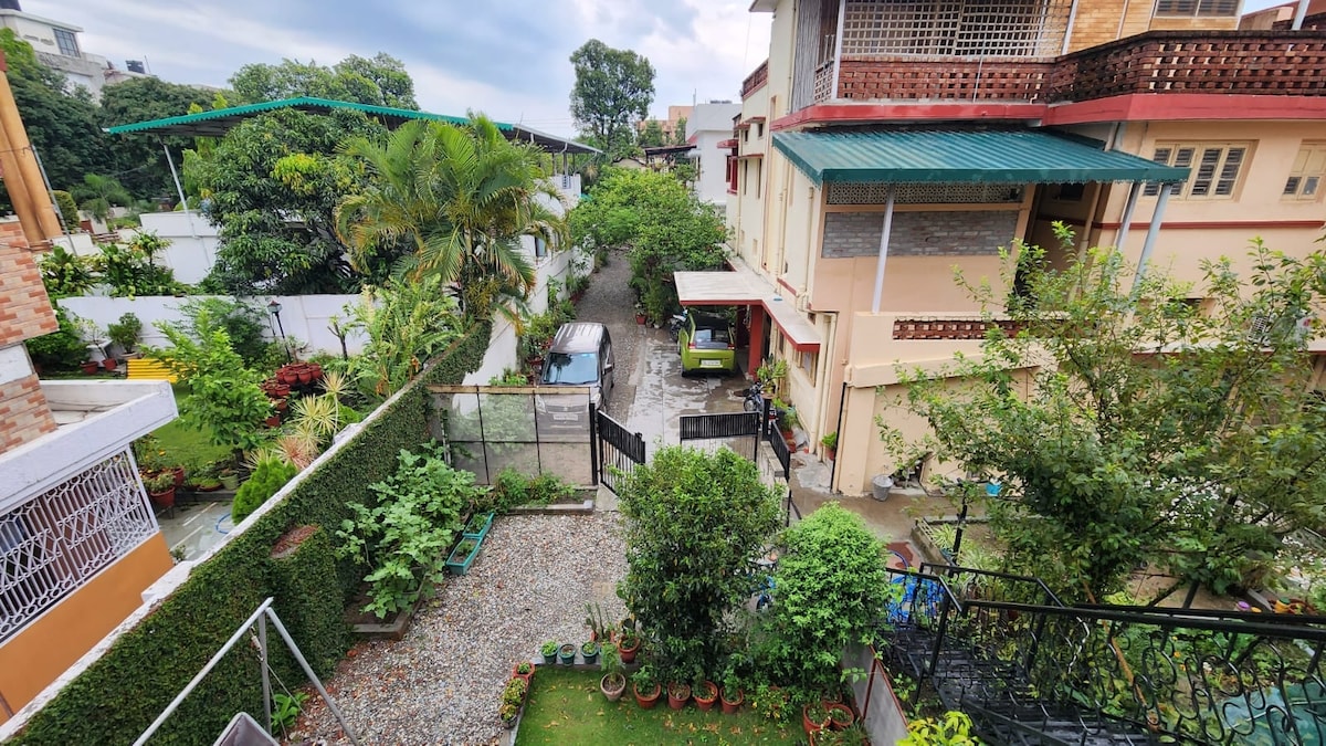 A garden path is visible, bordered by lush greenery and potted plants. The area features a gravel driveway with parked vehicles. Nearby buildings are partially visible, surrounded by trees and foliage, creating a serene and natural setting.