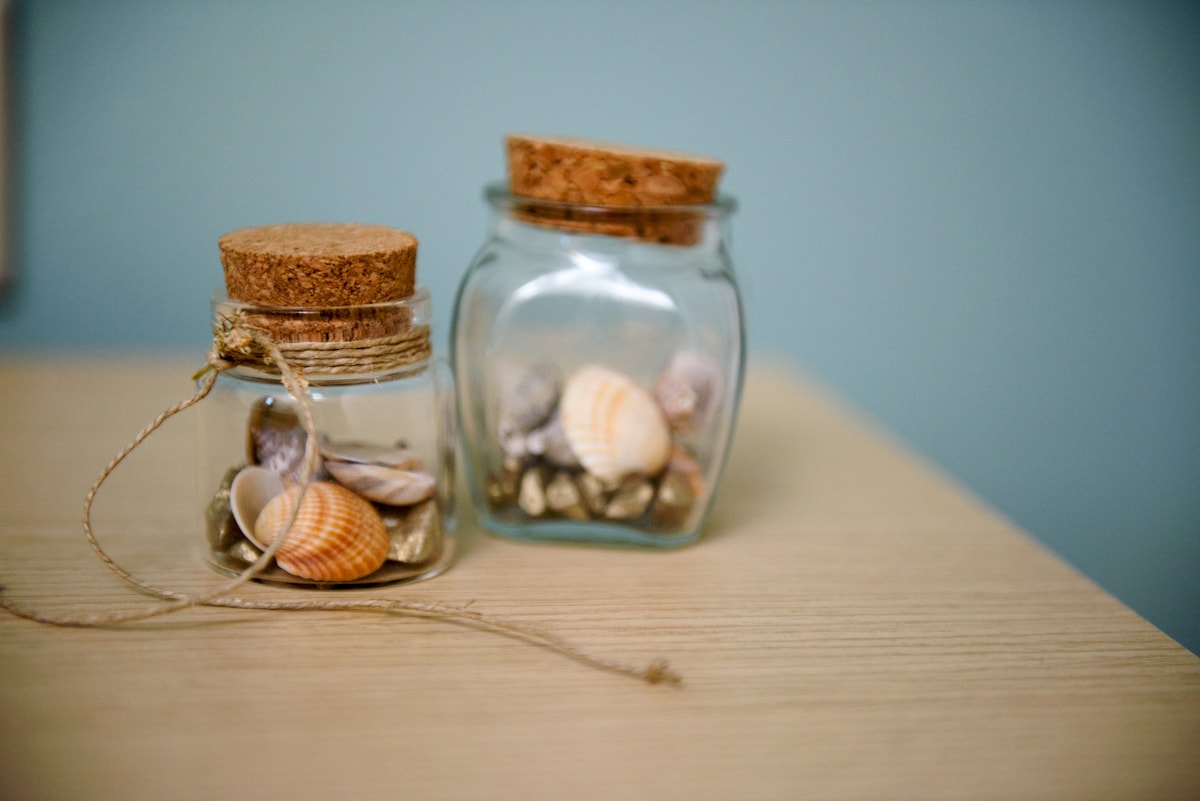 Two glass jars sit on a wooden table, each filled with a variety of seashells and small stones. The jars feature cork lids, and twine is wrapped around one of the jars, complementing the natural decor.