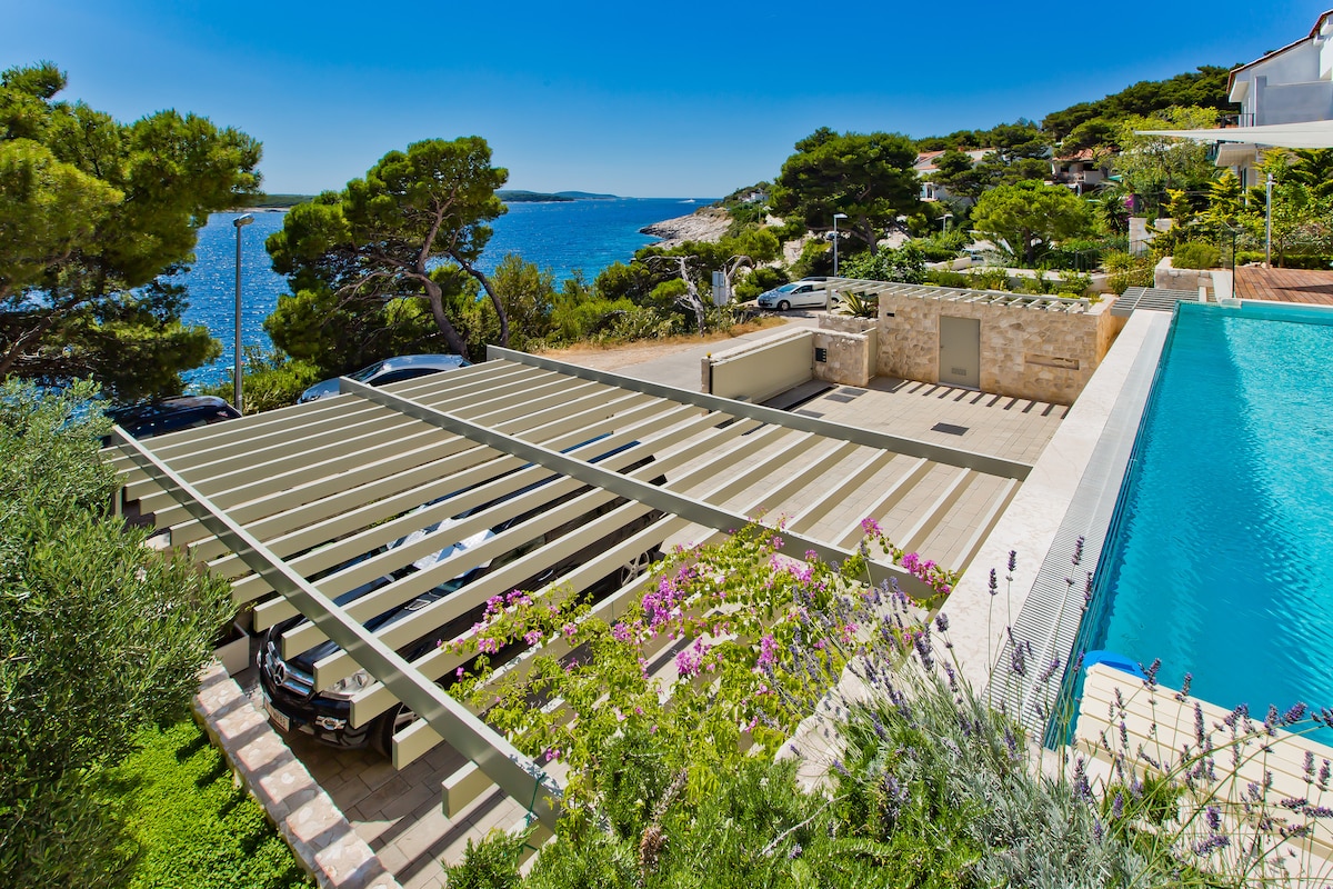 A serene view of the oceanfront property features a modern swimming pool alongside two parked cars under a spacious pergola. Lush greenery and flowering plants surround the area, enhancing the natural beauty of the coastal landscape, with the sea visible in the background.