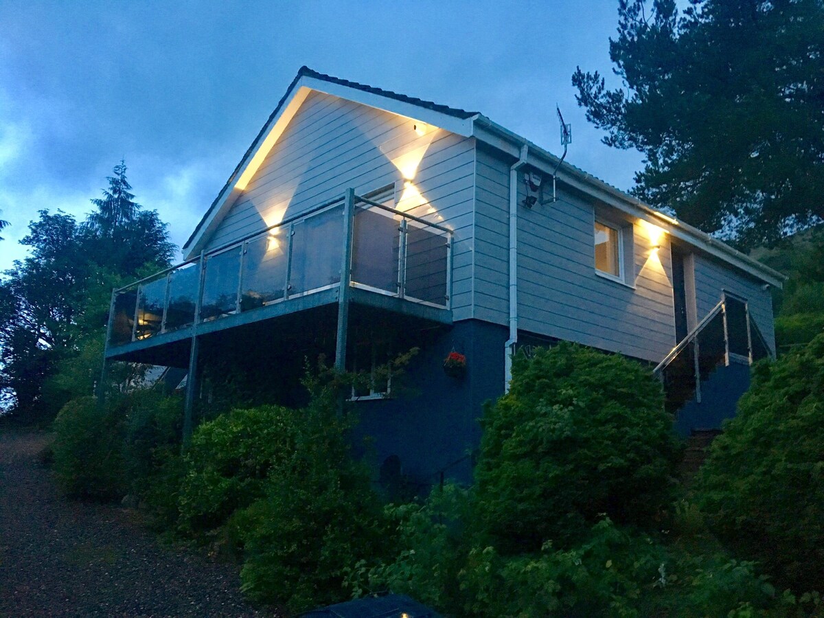 The exterior of the house is highlighted by soft lighting against a twilight sky. A spacious balcony is surrounded by landscaped greenery, with glass railings offering an unobstructed view. The structure features a sloped roof and is set amidst natural surroundings.