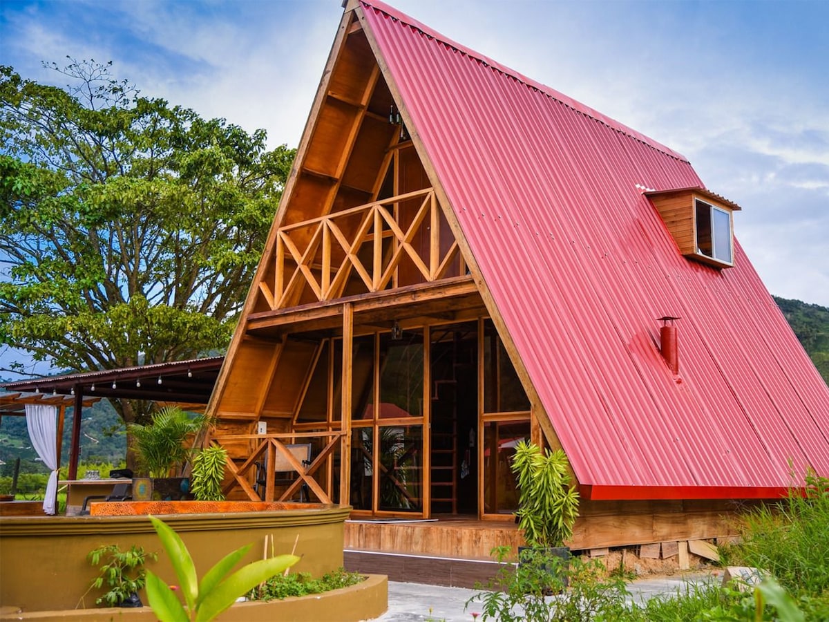 An A-frame cabin features a striking red roof and wooden façade, surrounded by lush greenery. Large glass windows provide views of the natural landscape, while a cozy outdoor seating area is visible, blending with the tranquil setting.