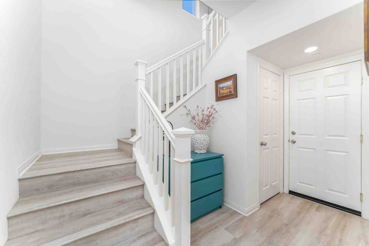 A bright entryway is highlighted by a staircase leading to the upper level. A small chest of drawers in a turquoise hue is positioned next to the door, with a decorative vase containing dried flowers on top. Natural light filters in, enhancing the spacious feel.