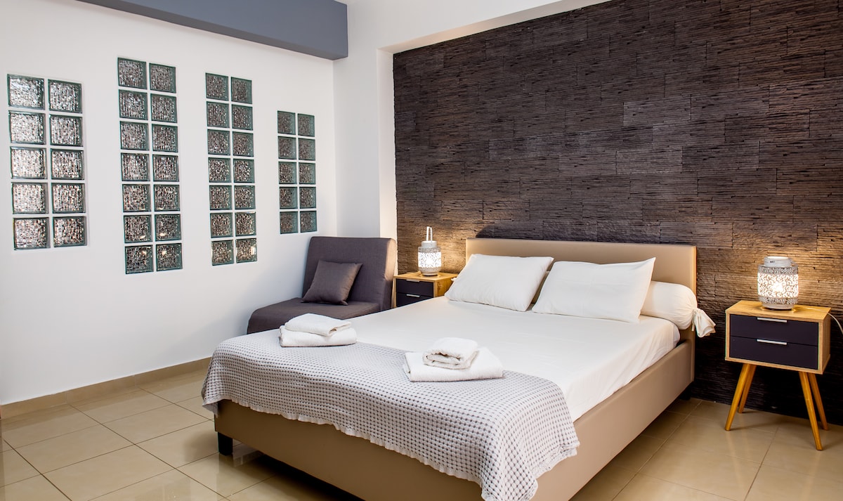 A spacious bedroom features a comfortable bed with neatly arranged linens and two side tables. A cozy lounge chair is positioned in the corner. Textured stone walls provide a modern backdrop, complemented by decorative glass panels that add visual interest.