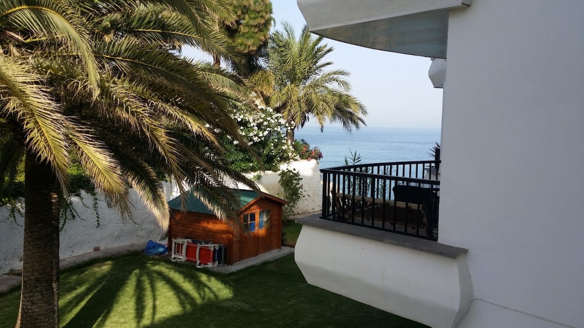 A view from the terrace showcases a well-maintained garden featuring a wooden shed and palm trees, with the sea visible in the background. The scene presents a tranquil outdoor space, framed by the white building's balcony railing.