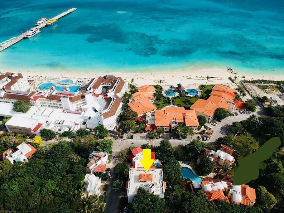 An aerial view captures the vibrant coastline of Playa del Carmen, with its clear azure waters and sandy beach. The surrounding area is dotted with lush greenery and charming buildings, showcasing a mix of accommodation and amenities within close proximity to the shore.