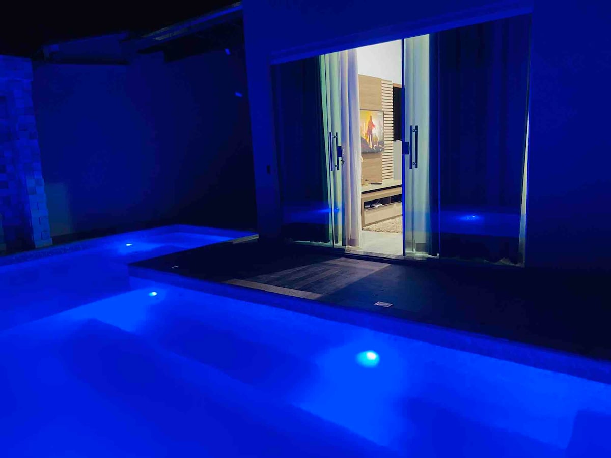 A serene pool area is enhanced by soft blue lighting reflecting off the water's surface. The view from the pool provides a glimpse of an indoor space through expansive glass doors, where a cozy seating area is visible.