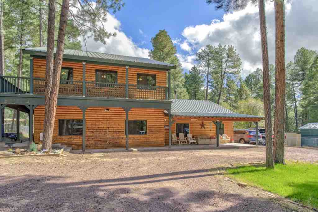 A large two-story cabin is framed by tall trees, featuring a wooden exterior and a shaded front porch with seating. The gravel driveway leads to the entrance, while green grass and natural landscaping create a welcoming outdoor environment.
