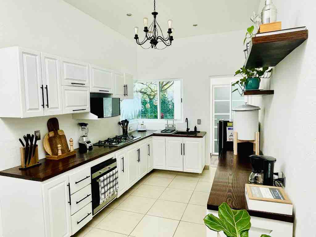 A well-equipped kitchen is showcased, featuring white cabinetry and a spacious countertop with wooden accents. A gas stove, sink, and various kitchen tools are visible. Natural light enters through a window, while decorative plants add a touch of greenery to the space.
