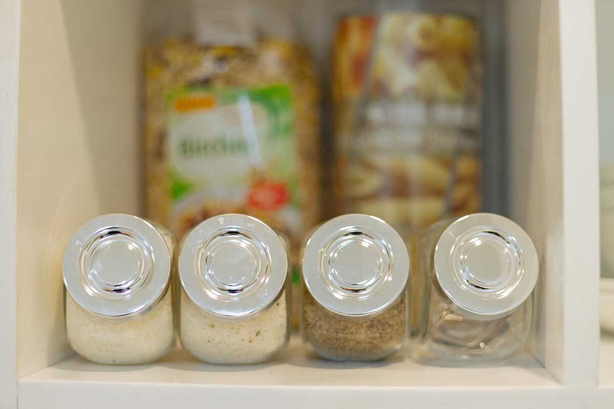 A collection of four glass jars, each with a silver lid, is displayed neatly on a white shelf. The jars contain various spices and ingredients, while a bag of dried food products is visible in the background.