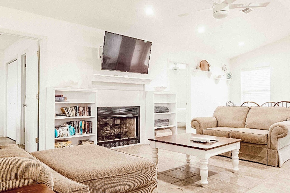 A light-filled living area features a comfortable sofa and armchair arranged around a central coffee table. A wall-mounted television is positioned above a white fireplace. Open shelving displays books and blankets, enhancing the welcoming ambiance of the space.