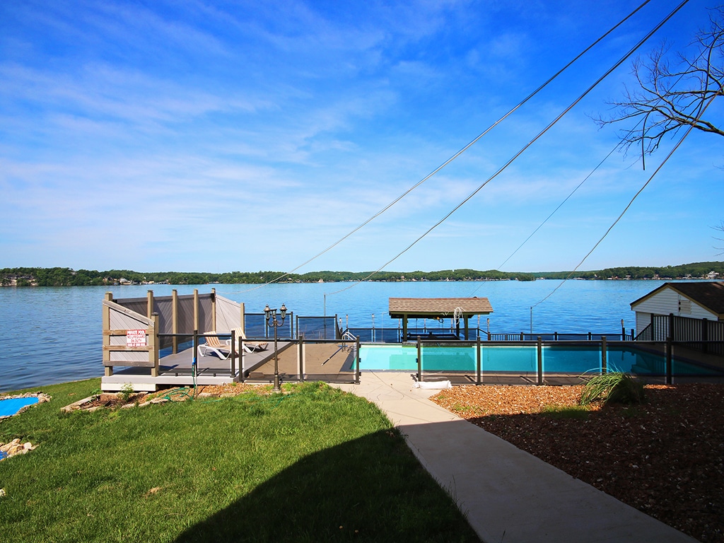 A view shows the serene lake with glassy water reflecting the blue sky. The foreground includes a grassy area, along with a saltwater pool bordered by a safety fence. A tiki bar and dock are visible near the water's edge, providing access and leisure space.