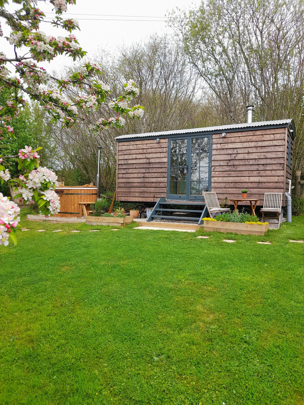 A shepherd's hut is nestled in a lush green garden surrounded by flowering apple trees. A wooden hot tub is visible nearby, with seating areas featuring wooden chairs and a small table. The outdoor space is well-kept, providing a serene ambiance.