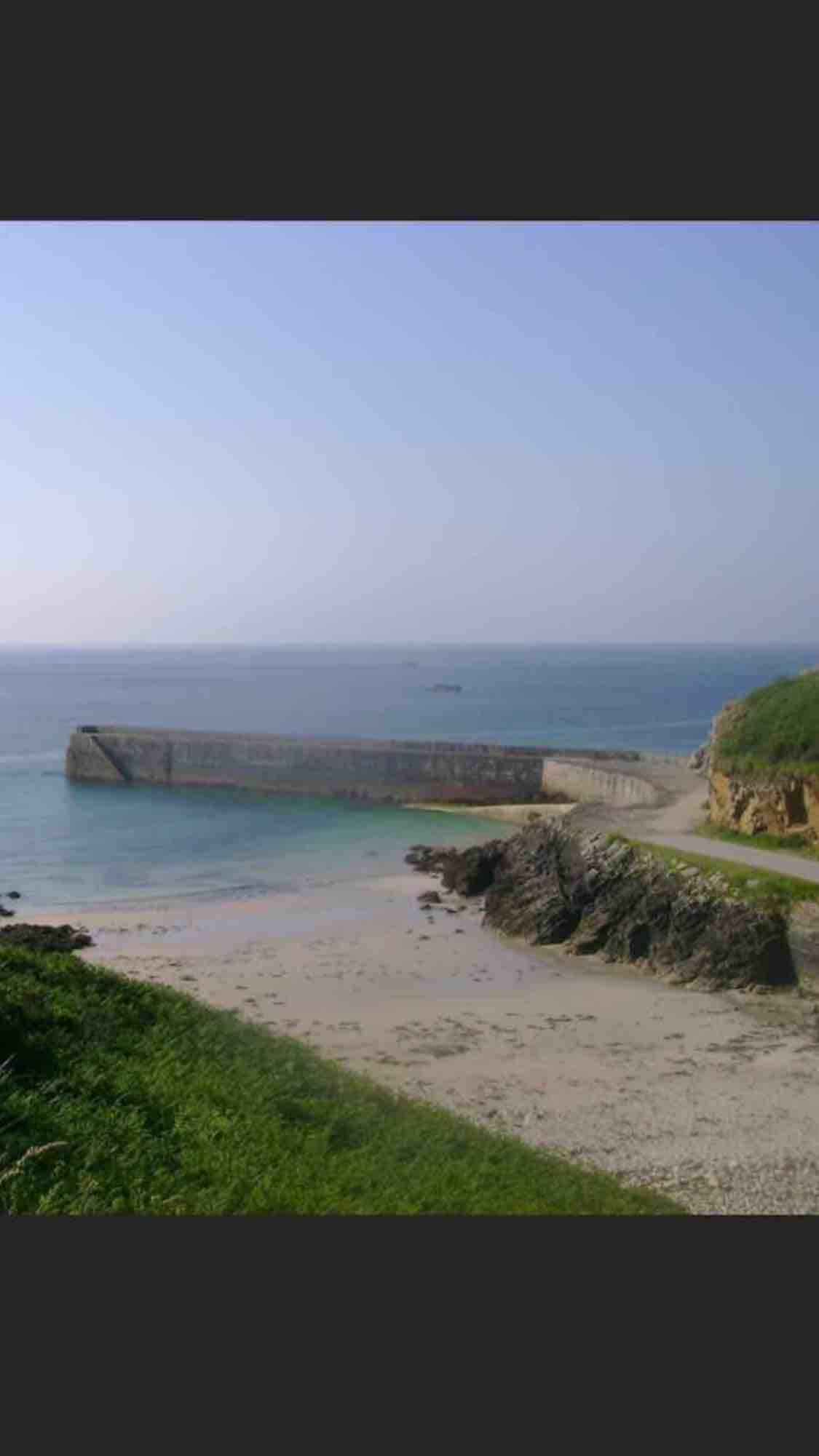 A serene coastal view is captured, showcasing a stone pier extending into calm waters. Gentle waves lap against the shore, bordered by rocky formations and patches of green vegetation. The sky appears clear with a subtle gradient of blue, enhancing the tranquil atmosphere of the scene.
