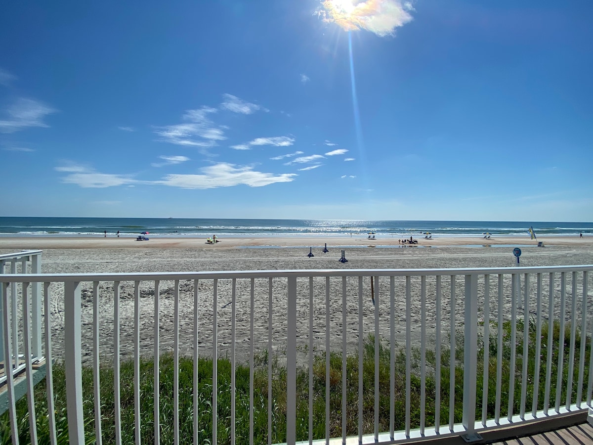 An oceanfront view showcases a sandy beach stretching toward the horizon, where the calm waves meet the sky. Guests can be seen relaxing under umbrellas, while a bright sun illuminates the scene, casting vibrant reflections on the water.