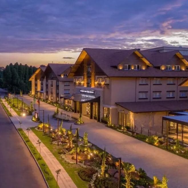An exterior view of the Wyndham Thermas Resort Spa showcases a charming structure with wooden accents and multiple gabled roofs. Warm lights illuminate the entrance and landscaped pathways, creating a welcoming atmosphere against a twilight sky.