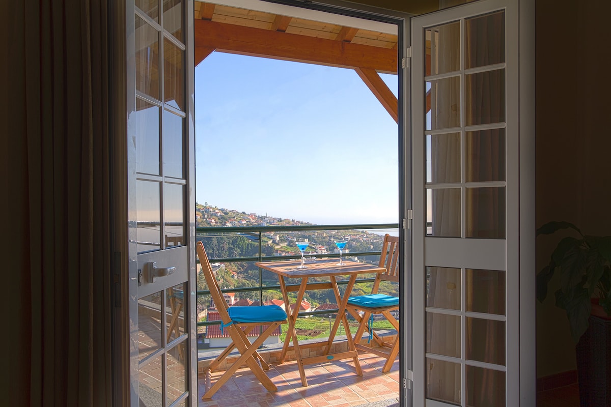 A balcony is framed by open French doors, featuring a wooden table with blue-topped chairs. The view expands over the landscape, highlighting the gentle hills of Madeira under a clear sky.