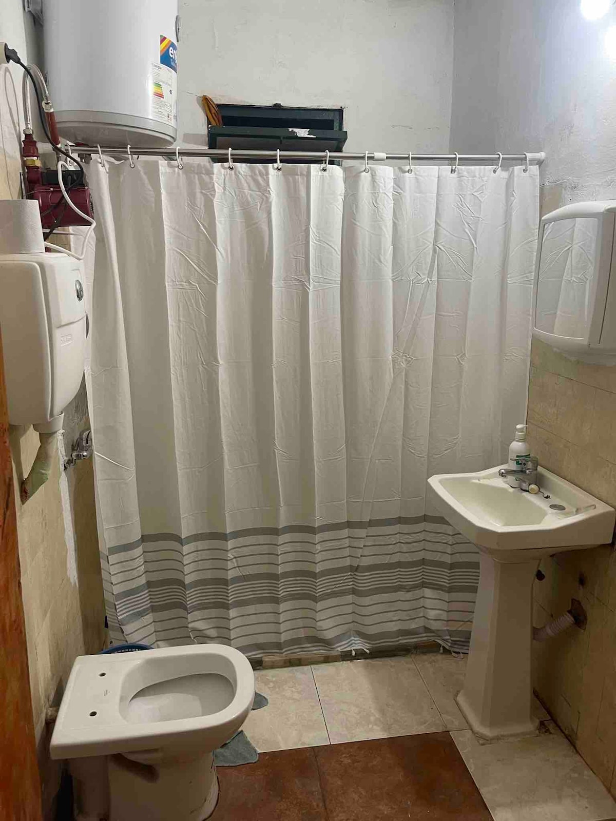 A functional bathroom is presented, featuring a shower area with a white curtain, a pedestal sink with a faucet, and a toilet. Wall-mounted dispensers are visible, providing convenient access to toiletries. The flooring is composed of contrasting tiles, enhancing the overall practicality of the space.